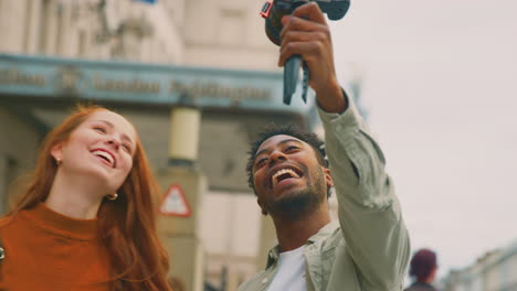 Pareja-Joven-Viajando-Juntos-Por-La-Ciudad-Haciendo-Vlogging-Con-Una-Cámara-De-Video-En-Un-Trípode-Portátil