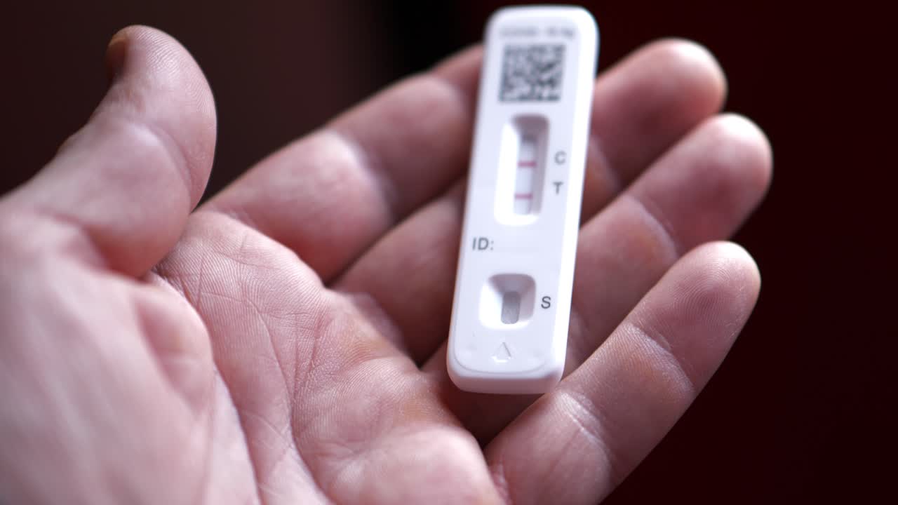 白人男性の開いた指の手にある、コントロールとテストに2本の赤い線が表示されたCOVID-19陽性抗原検査キット。カメラがゆっくりとパンし、彼が検査キットをわずかに調整する。