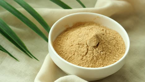 close-up view of a bowl of light brown powder on a beige fabric background with a green leaf