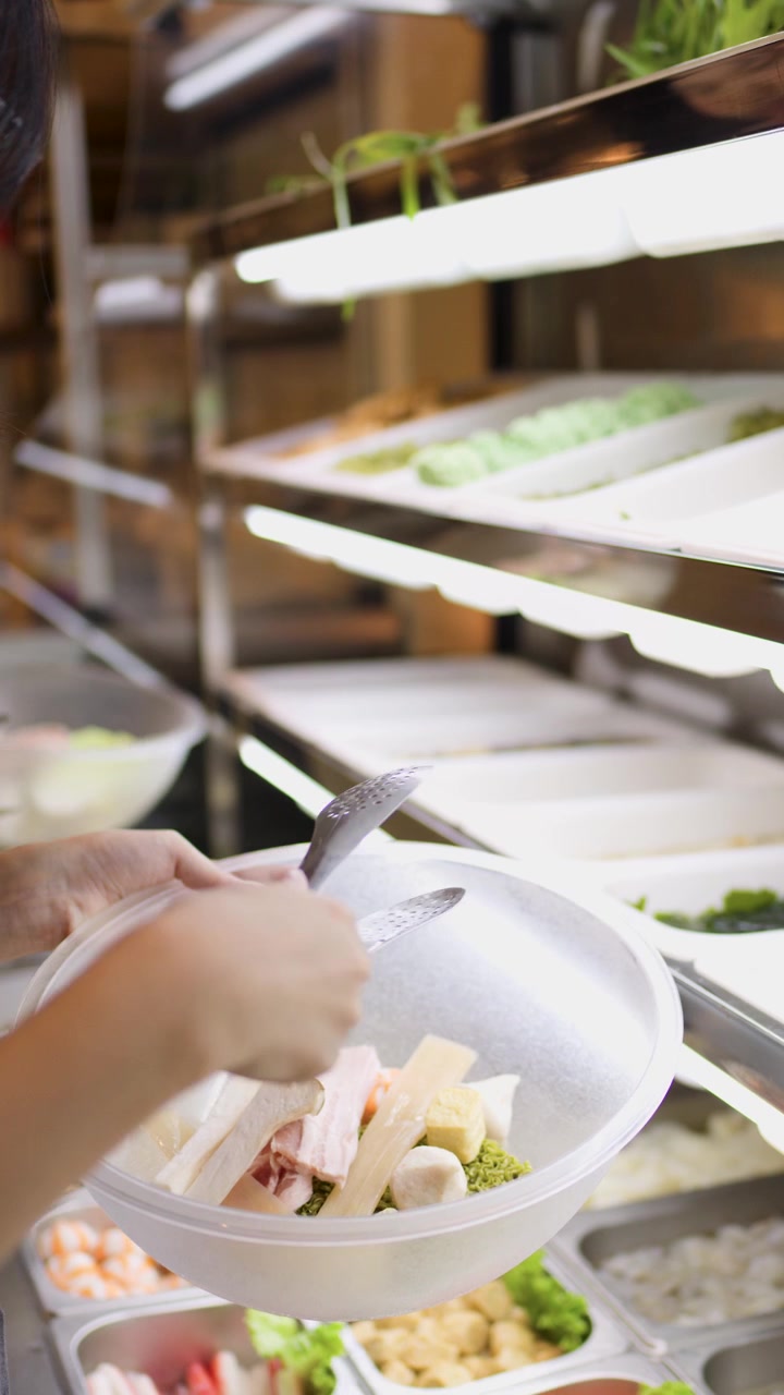 Una persona riempie una ciotola con verdure miste e noodles a un bancone buffet self-service al coperto.