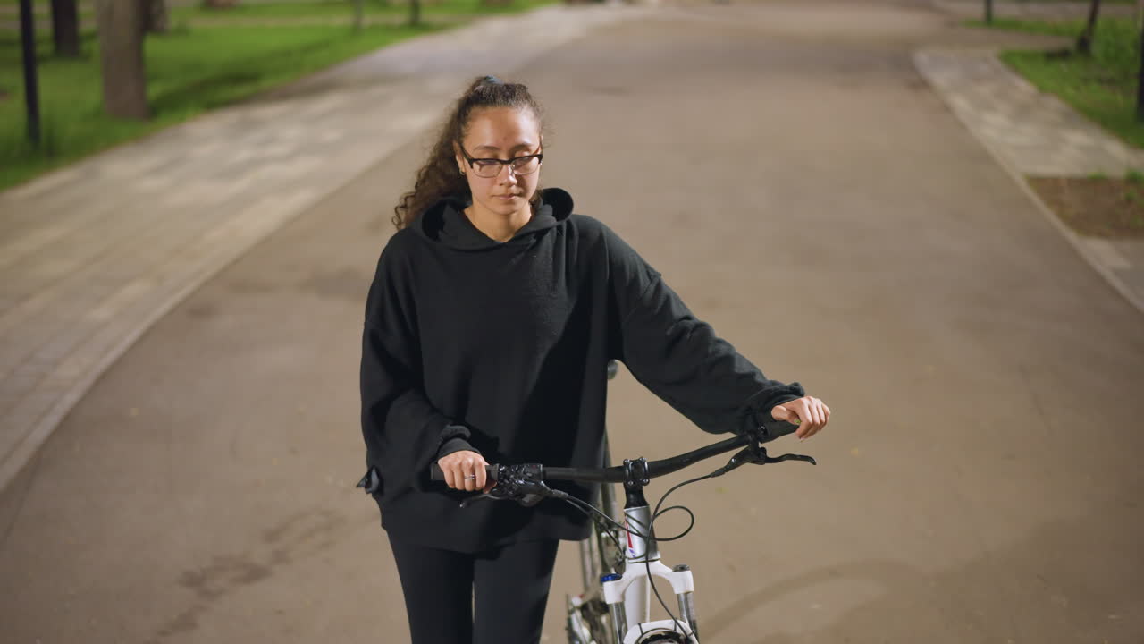 Woman Stabilizes Bike Quietly At Night, Woman Balances Her Bicycle Under Soft Street Lighting, Serene Nighttime In Park Where Woman Steadies Her Bicycle Among Trees And Warm Streetlights