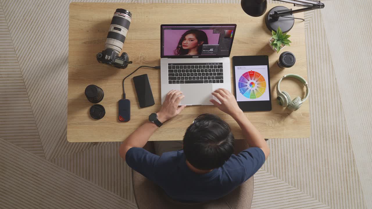 vista superior de un editor masculino mirando los esquemas de colores en una tableta mientras está sentado en el espacio de trabajo usando una computadora portátil al lado de la cámara editando la foto de una mujer en casa
