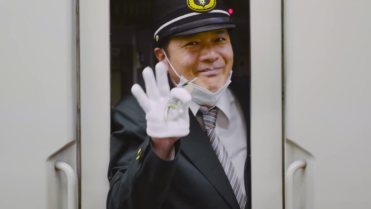 Japanese train conductor blows whistle, points forward and waves to camera while smiling