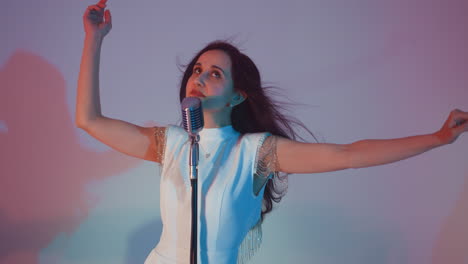 singer in a white gown with eyes closed, moving hands gracefully in the air while performing into a vintage microphone. set against a soft gradient background of blue and pink