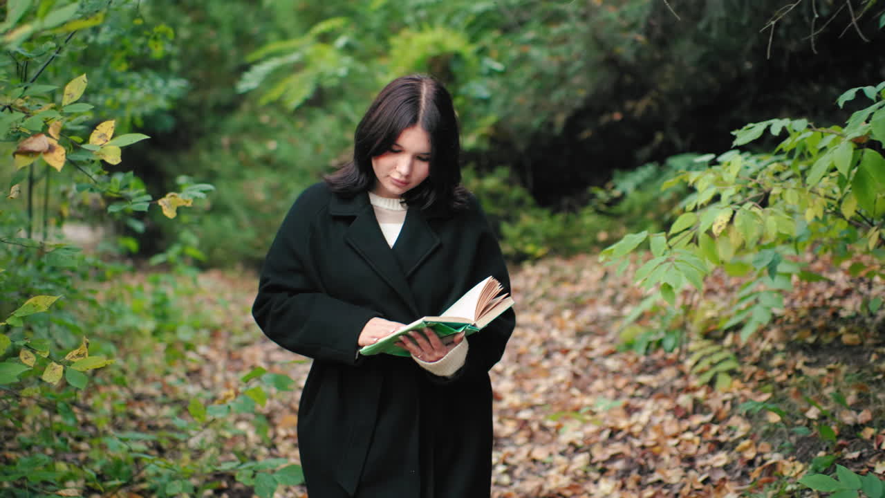 Leg view shows graceful woman in autumn coat walking forest path, black boots crunching dry leaves while browsing book and looking around, calm pace, soft bokeh greenery, quiet travel mood