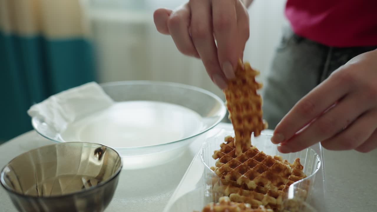 플라스틱 용기에서 와플을 집는 손, 금속 볼이 근처에 있는 주방 조리대에서 클로즈업한 아침 간식 의식; 빨간 셔츠를 입은 사람이 손가락으로 바삭한 황금빛 와플을 떼어내는 모습, 촉각적인 이미지