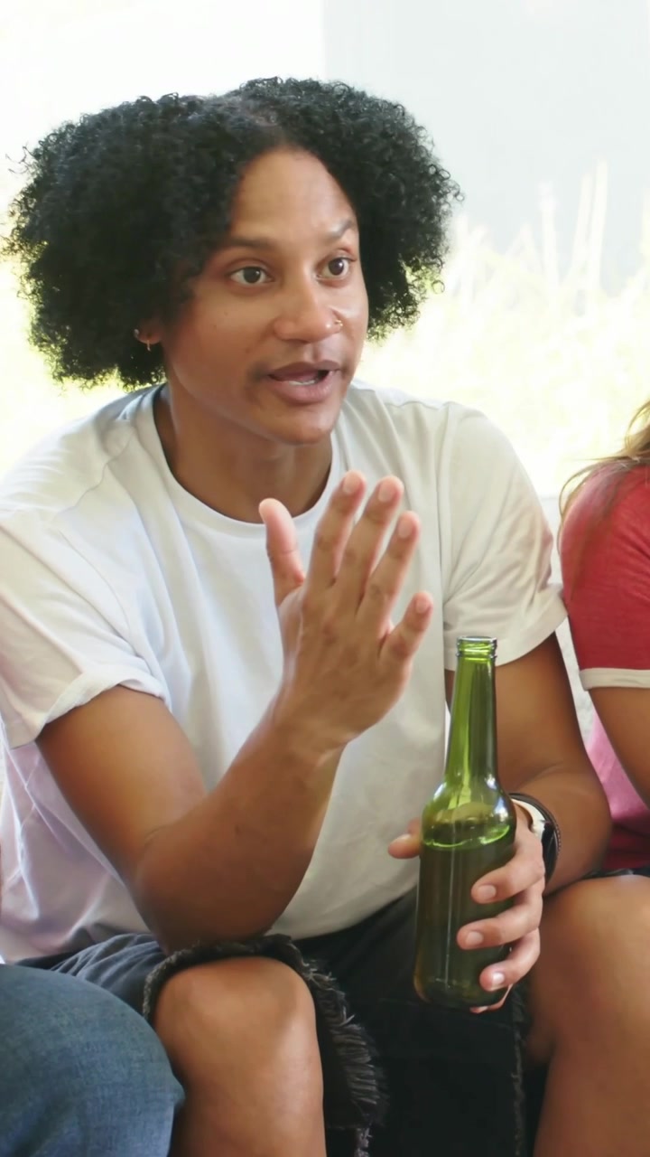 Vertical video: During chat man in white tee emphasizing point on couch, holding green bottle