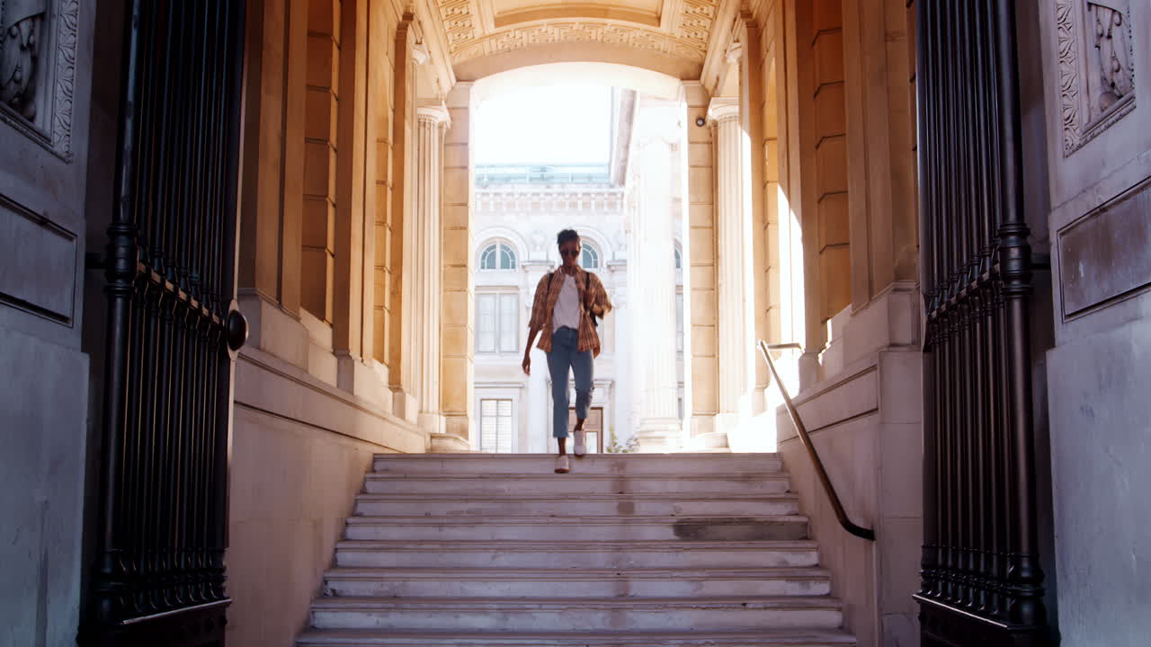 una joven negra con una camisa a cuadros y pantalones vaqueros azules caminando por las escaleras vistas a través de la puerta de entrada fuera de un edificio histórico, vista frontal
