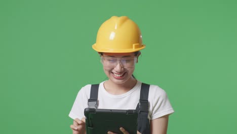 close up de una trabajadora asiática que usa gafas de protección y casco de seguridad sonriendo y usando una tableta mientras está de pie en el estudio de fondo de la pantalla verde