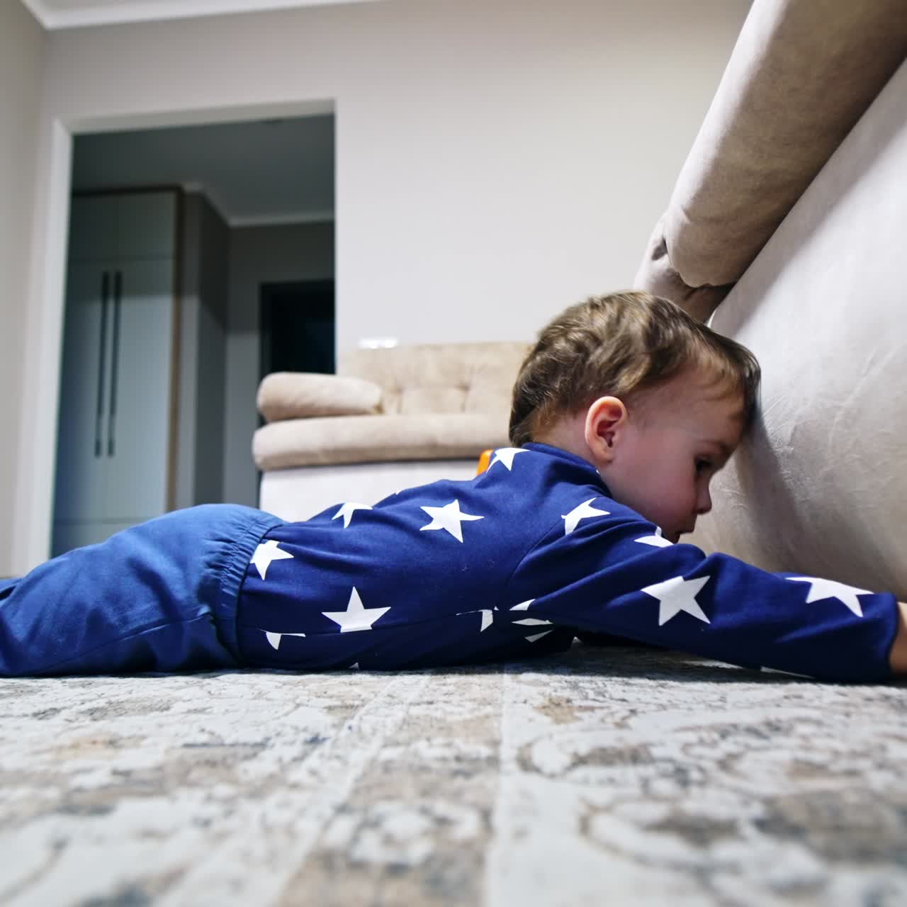 Toddler boy lies on the floor reaching his hand under the sofa. Baby wants to get something from under the couch