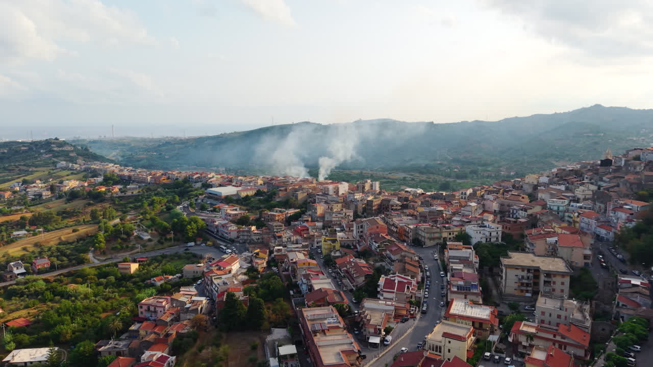 Villaggio siciliano in Italia con tetti rossi circondato da verdi colline, vista drone, fumo in lontananza, atmosfera rurale tranquilla.