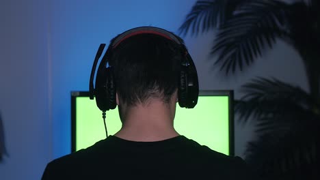 gamer puts his headset with a mic on and starts playing computer games on green screen monitor