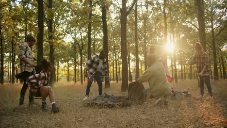 A-happy-team-of-six-people-on-a-hike-arrange-their-things-and-set-up-a-tent-to-spend-the-night-in-a-summer-green-forest-with-withered-grass.-A-break-during-a-group-hike-in-the-summer-forest