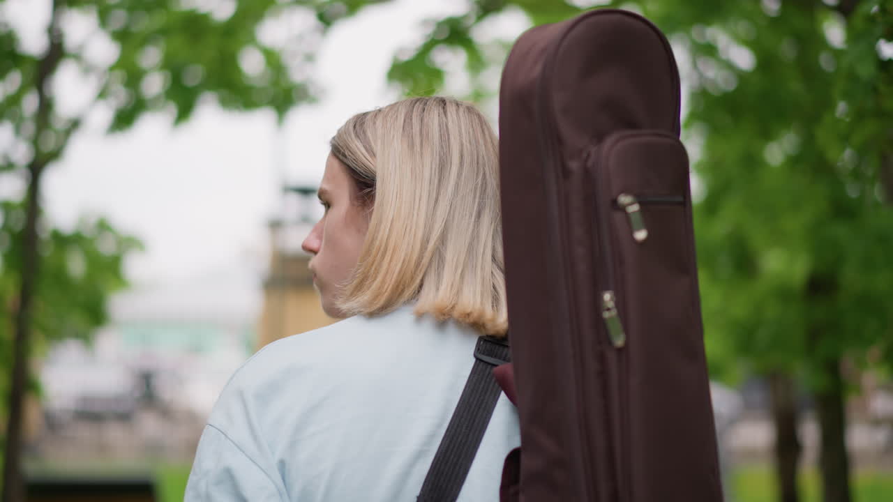 Un chitarrista bianco con custodia per chitarra cammina in un parco urbano alberato, con la custodia imbottita sulla schiena, giacca di jeans e fascia per capelli, espressione pensierosa, profondità di campo ridotta, punto di riferimento della città e lampione