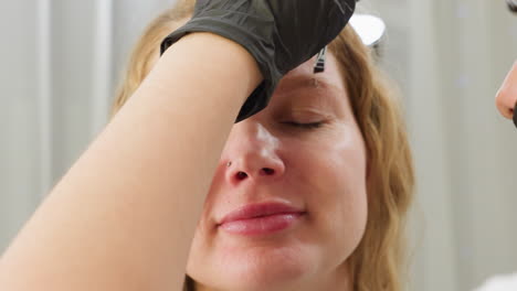 smiling client wearing nose ring relaxes as beautician applies cream to eyebrow during cosmetic session and beautician gloved hand gently works while client enjoys professional salon service