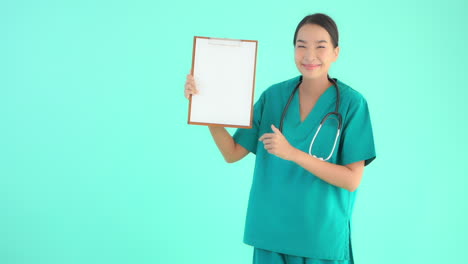 young asian model in medical scrubs with stethoscope holds up clipboard with blank white page, smiles and gives thumbs up