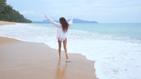 With-her-back-to-the-camera,-a-healthy,-fit,-woman-wearing-a-bathing-suit-and-a-white-shirt-coverup-runs-along-the-surf-as-it-rushes-to-the-shore