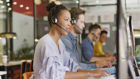 Happy-diverse-business-people-sitting-at-table-and-using-phone-headsets-at-office