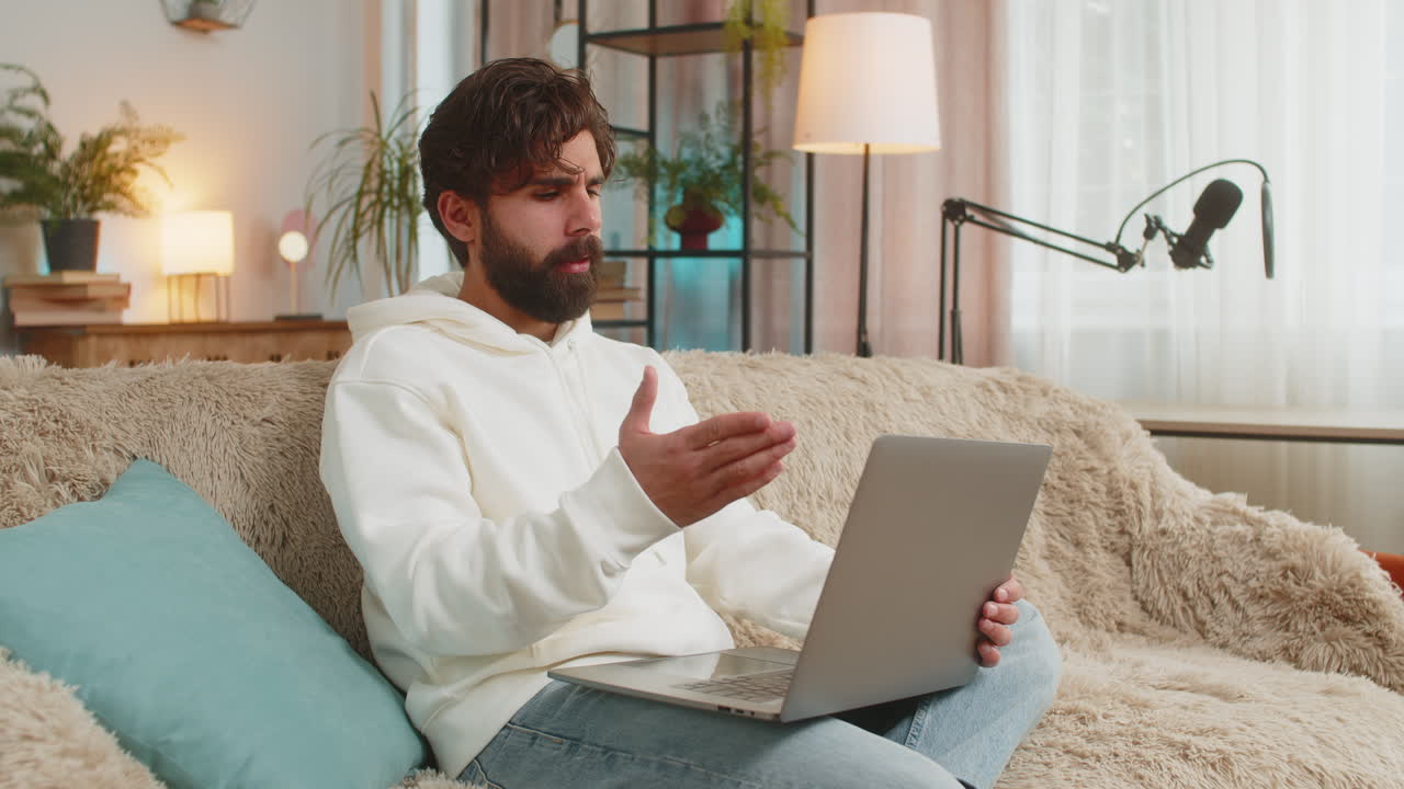 Upset young indian man freelancer using laptop engages in heated conversations sitting on sofa