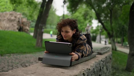 happy girl in a plaid shirt lies on a bench and moves her legs while watching lessons on a tablet in the park