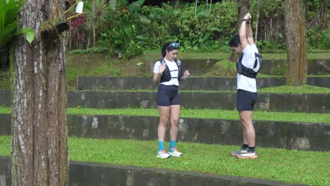 an asian couple puts drink pouches into their vest pockets before starting their workout in a tree-filled park