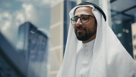 portrait of a successful happy muslim businessman in traditional white kandura wearing glasses riding glass elevator to office in modern business center. saudi, emirati, arab businessman concept.