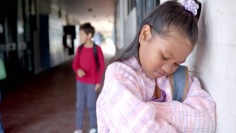 in school, girl leaning against wall looking sad, boy in background with backpack