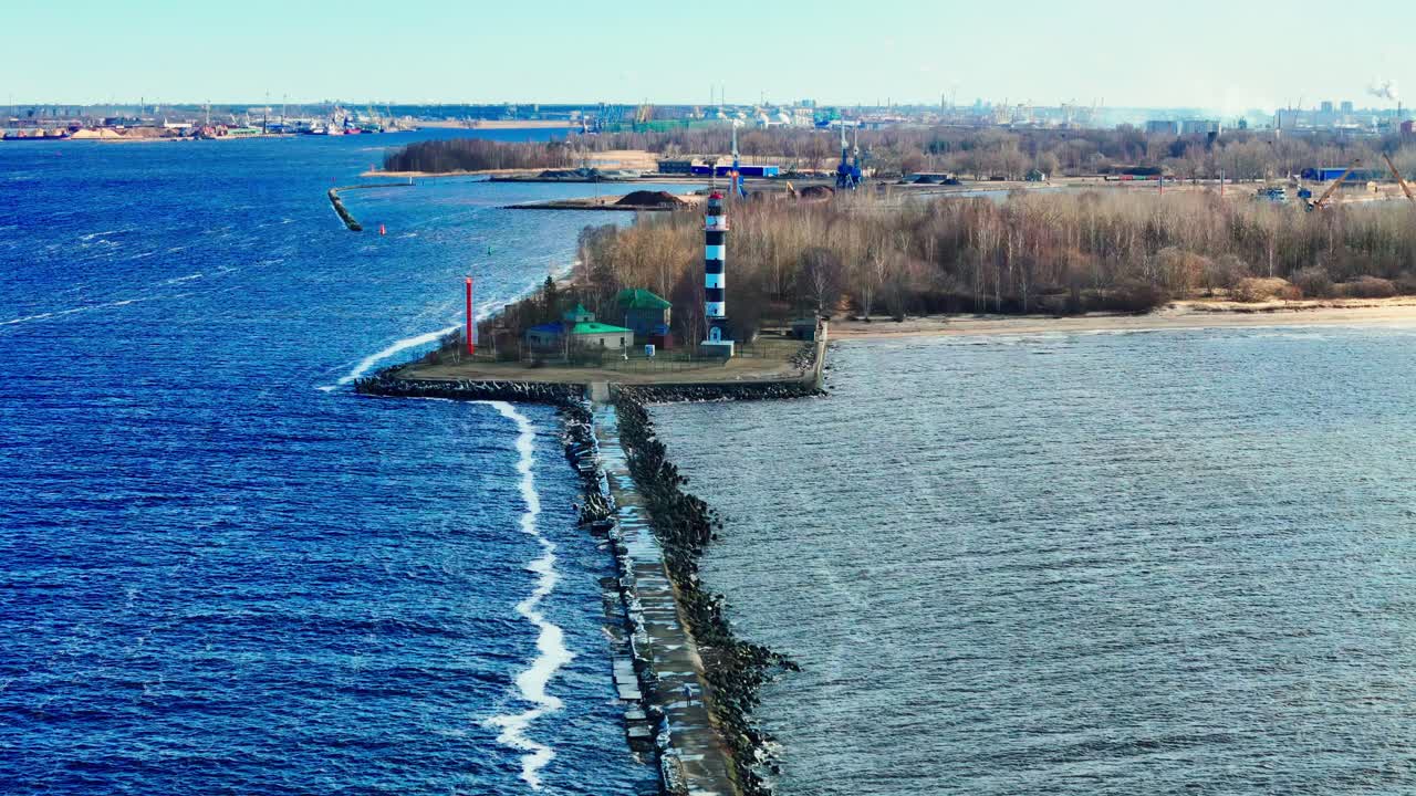 Aerial view of a black and white lighthouse on a slender stone jetty, separating vibrant blue sea from tranquil estuary, bordered by forested coastline and distant city.