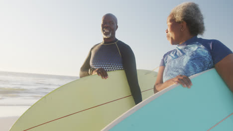 Happy-senior-african-american-couple-walking-and-holding-surfboards-at-beach,-in-slow-motion