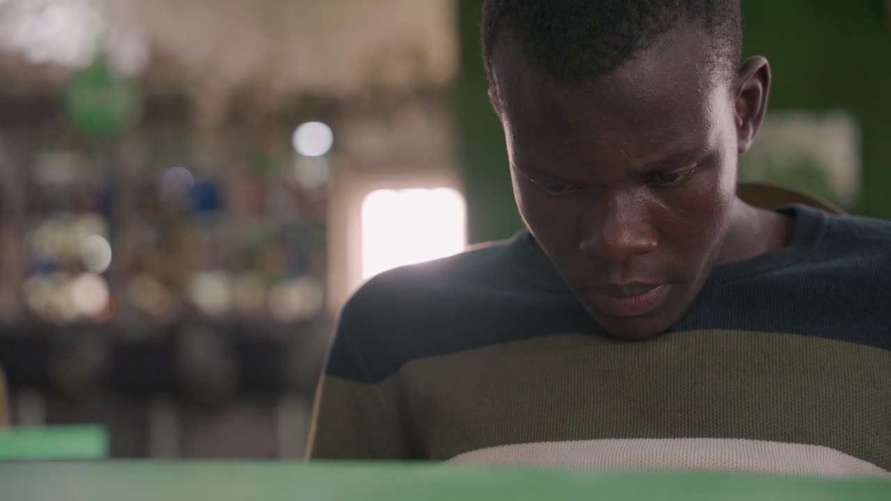 Black man reading love note in cozy cafe, soft film grain, portrait of nervous anticipation before date, downcast gaze, blurred bar shelves in background, warm amber light, intimate romantic mood