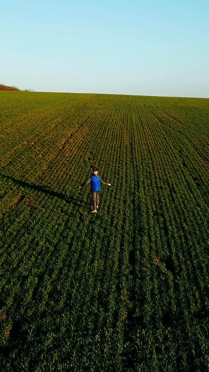 Cute boy running and turning around on the spacious green field in spring. Little kid in sportsuit playing outdoors on the natural background. Aerial view Vertical video