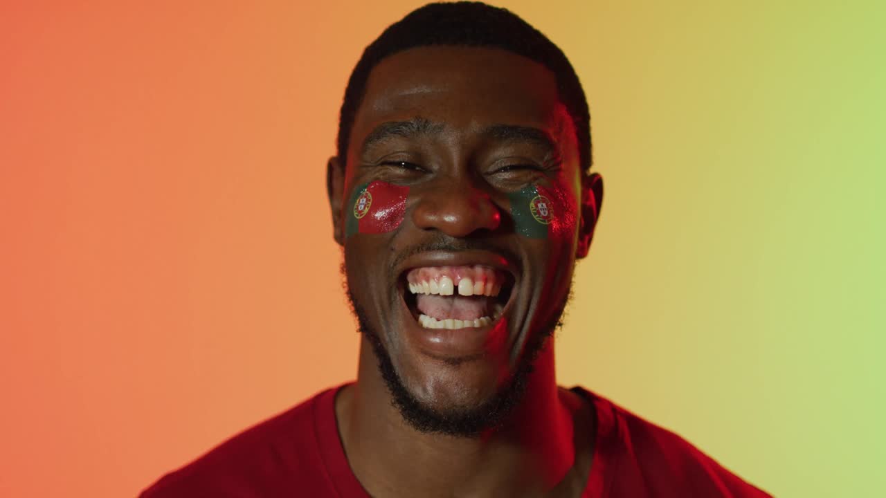 портрет счастливого афроамериканского мужского болельщика с флагом португалии на щеках над розовым освещением