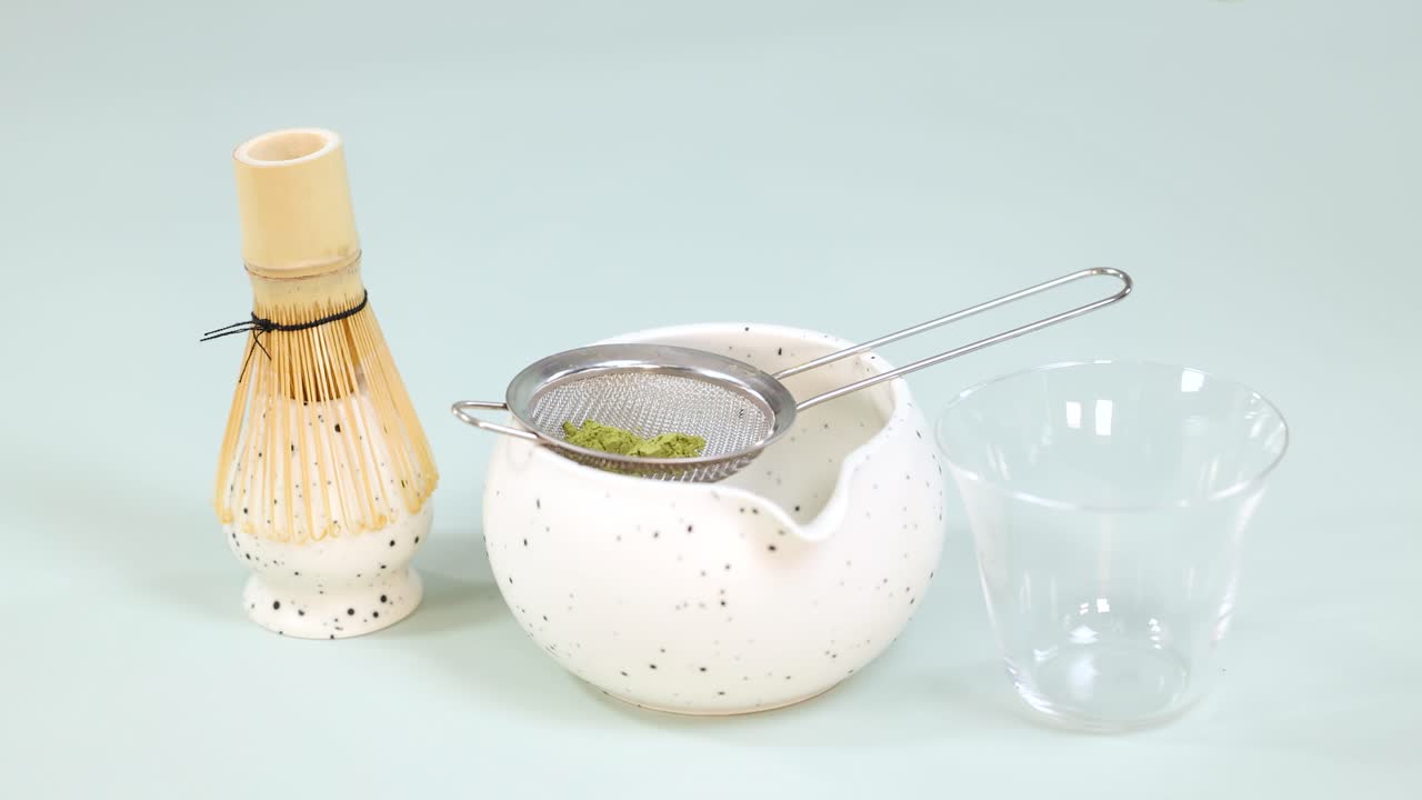 Hand sifts matcha powder into bowl using bamboo scoop, strainer, under soft studio lighting
