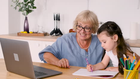 little girl drawing sitting at table in kitchen with her happy grandmother while having a video call on modern laptop 1