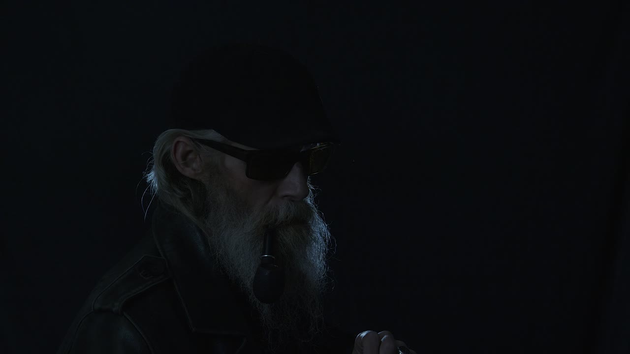 Bearded man in flat cap brings lit match to pipe, backlit in dark room