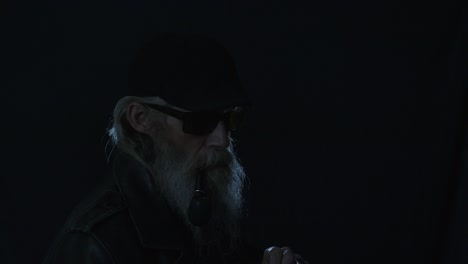 bearded man in flat cap brings lit match to pipe, backlit in dark room