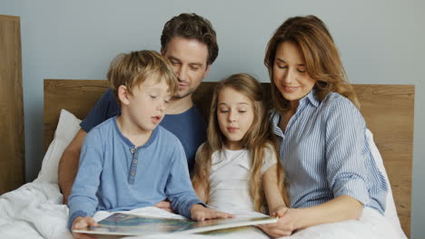 madre y padre jóvenes sentados en la cama por la mañana con su pequeño hijo y su hija, están leyendo un libro interesante