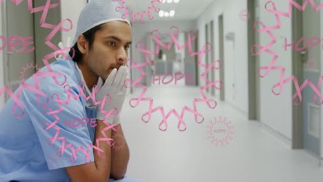 Animation-of-pink-globe-and-people-logos,-over-distressed-male-surgeon-in-hospital-corridor