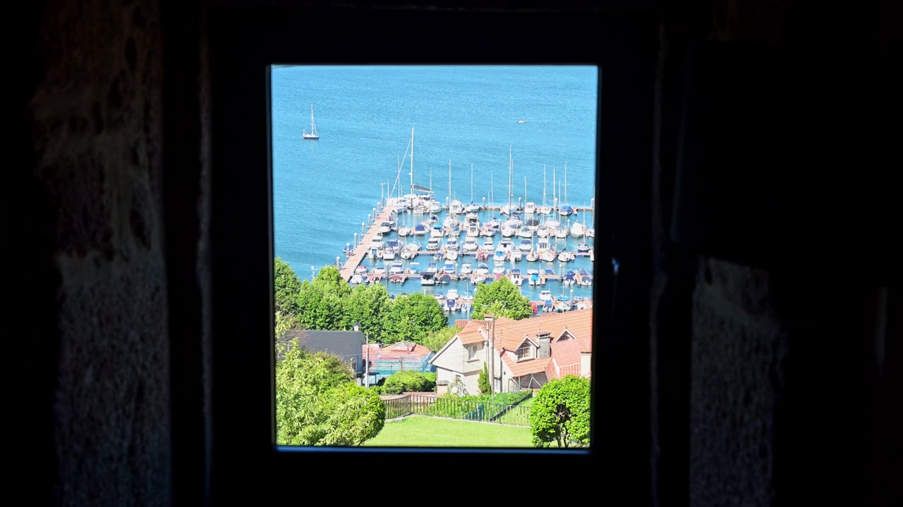 View from a dark room looking through a window frame at the Vigo estuary marina in Galicia, Spain. Documents coastal real estate perspective, architectural framing POV, and Atlantic lifestyle.