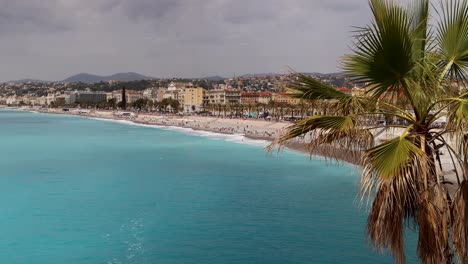 Promenade-des-Anglais-and-beach-in-Nice,-France,-with-turquoise-water-and-palm-tree-on-cloudy-day