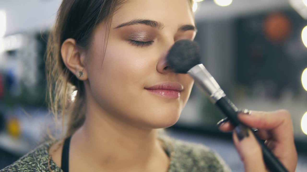 Close Up view of professional makeup artist's hands applying facial blusher on young woman's skin using special brush. Slow Motion