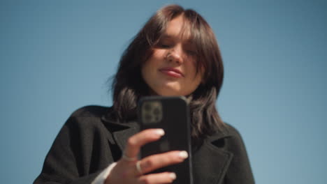 mujer alegre sonriendo, con el viento revoloteando su cabello, centrada en su teléfono, disfruta del momento al aire libre en un parque con un cielo azul claro