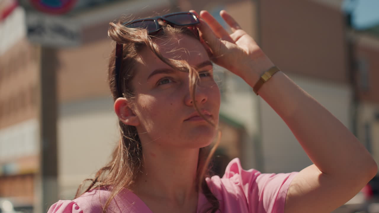 Calle soleada, mujer caucásica con smartphone y vestido rosa, gafas de sol, ajustándose el pelo, mirada pensativa, ambiente de bloguera de moda, tonos cálidos, edificios de ladrillo en el centro, calor veraniego, primer plano natural