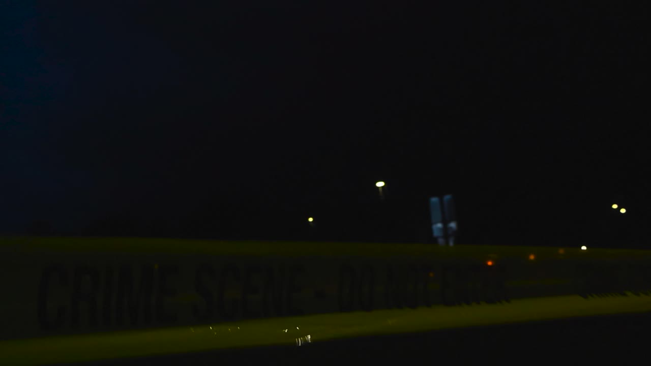 Close up view of a yellow colored police forensics investigation Crime Scene tape pulled in front of a dark skatepark area during a rainy night with bokeh street lights backlighting the ribbon dimly