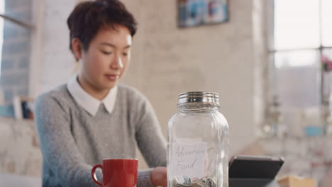 young asian woman putting money coins into savings jar at home