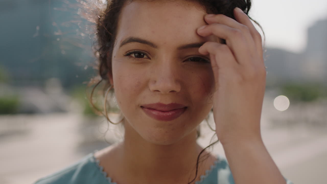 retrato en primer plano de una joven hispana linda que mira con confianza a la cámara corriendo de la mano a través del cabello con un fondo urbano soleado
