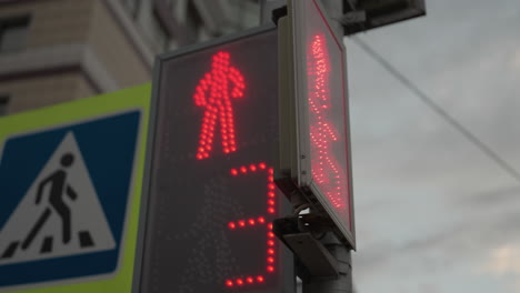 close-up view of a digital pedestrian traffic light displaying a red signal and countdown timer, the traffic light is positioned beside a pedestrian crossing sign, with a city building