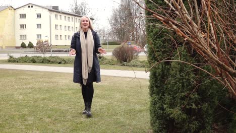 a woman with long blonde hair smiles and speaks while standing outdoors in front of evergreen trees, dressed warmly in a navy coat and beige scarf.