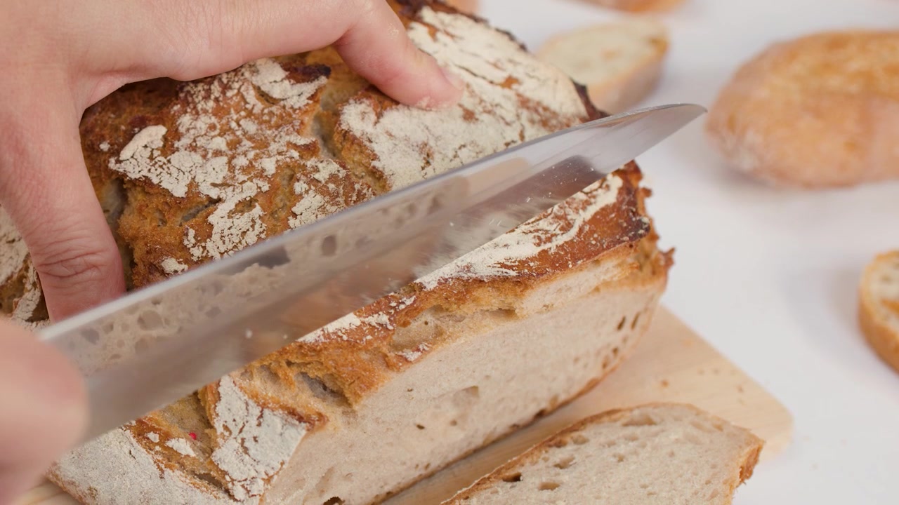 Primo piano di mani che usano un coltello per affettare pane artigianale a lievitazione naturale su superficie bianca