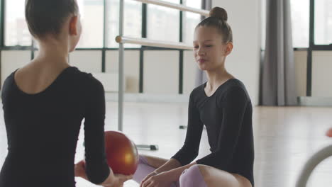 Zwei-Gymnastische-Blonde-Mädchen-Unterhalten-Sich,-Während-Sie-Mit-Einem-Ball-Spielen,-Der-Auf-Dem-Boden-Sitzt,-Bevor-Sie-Mit-Der-Ballettklasse-1-Beginnen